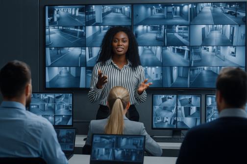 A woman presents confidently in front of a video wall displaying multiple security camera feeds. Three colleagues sit, watching attentively with laptops.