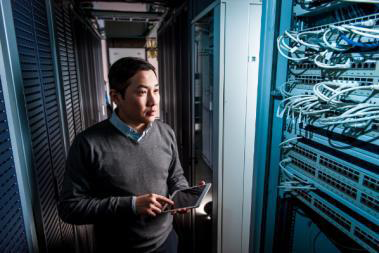 A person stands with a tablet in a dimly lit server room, surrounded by server racks and tangled cables. The scene conveys focus and technological expertise.